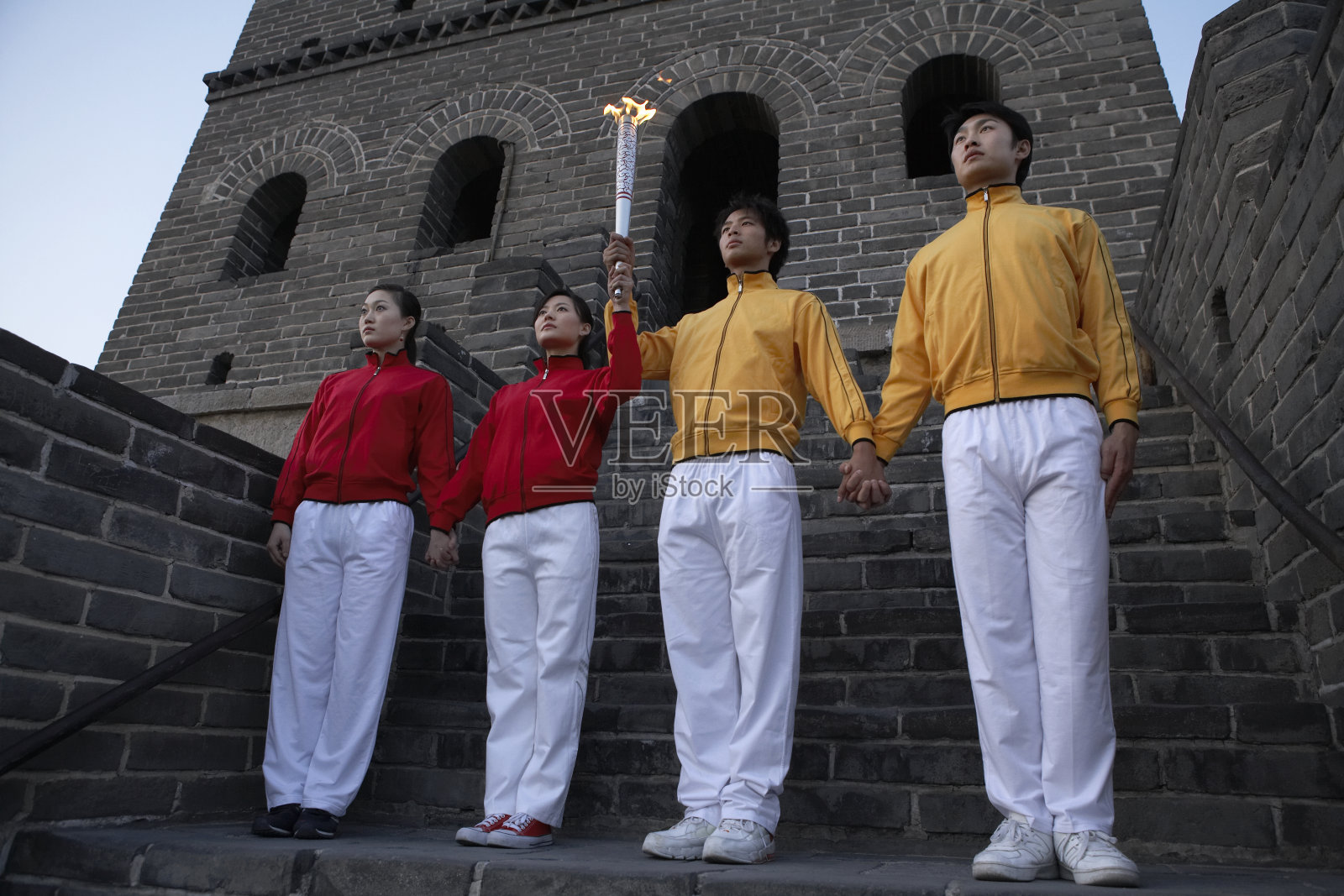 四位身穿运动服的年轻人手牵手的站在长城烽火台前，举着一支火炬照片摄影图片