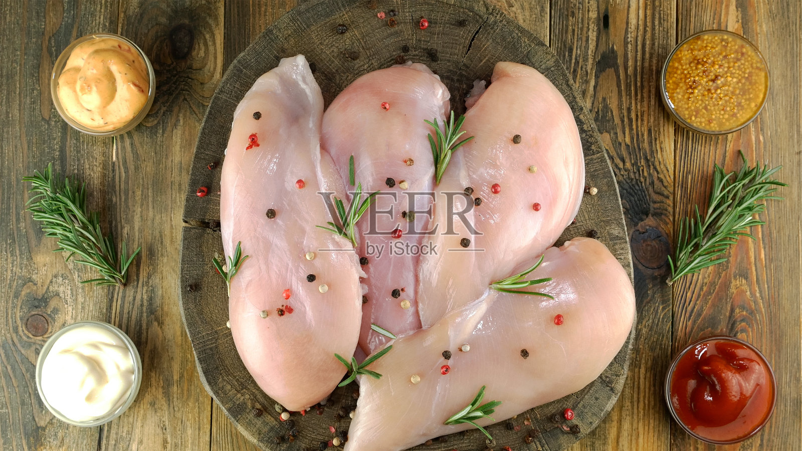 新鲜的生鸡胸肉，撒上胡椒粒，迷迭香和调味汁，放在圆木板上。照片摄影图片