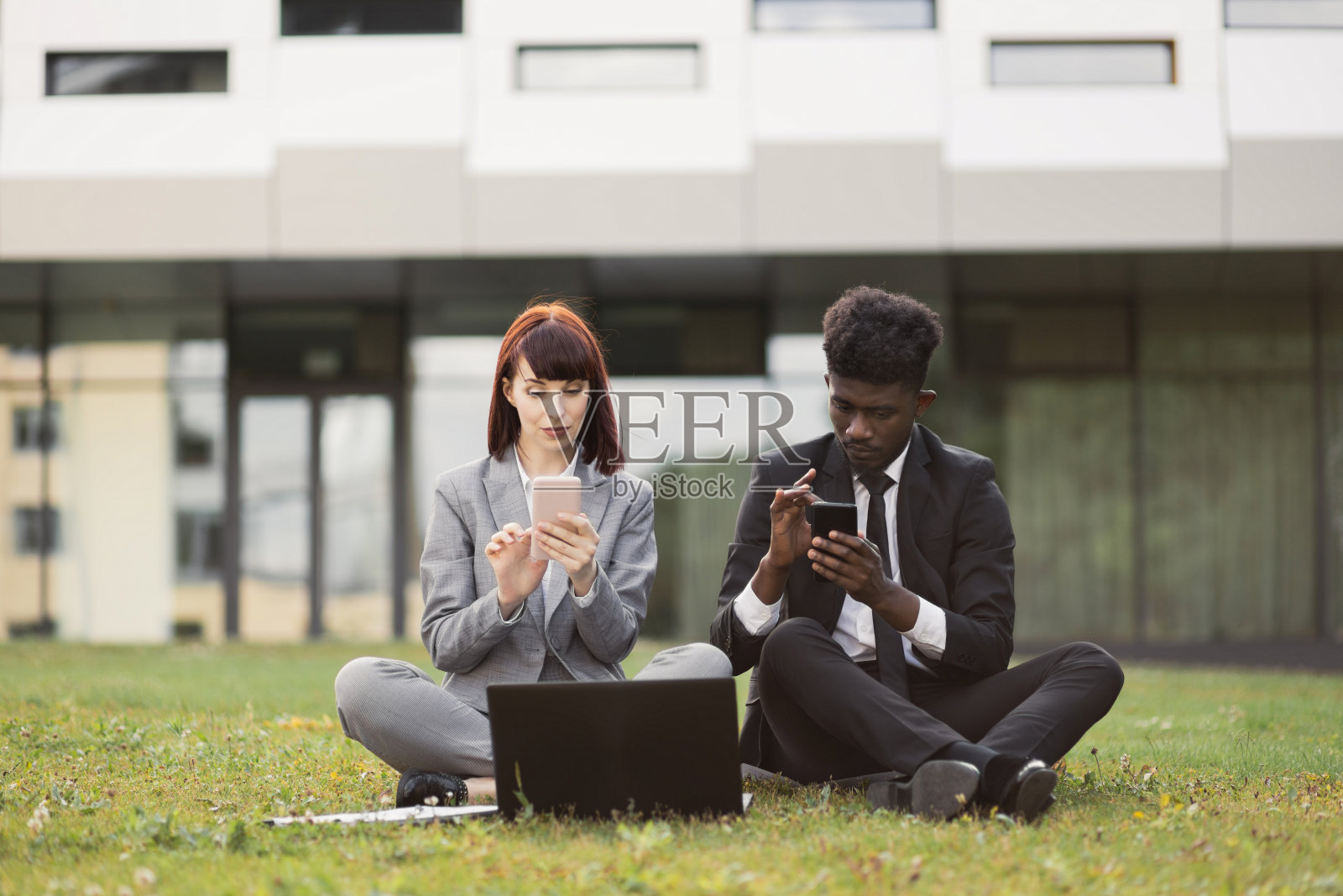 多民族的商务同事，享受户外休息，坐在办公室附近的草地上，使用电话照片摄影图片