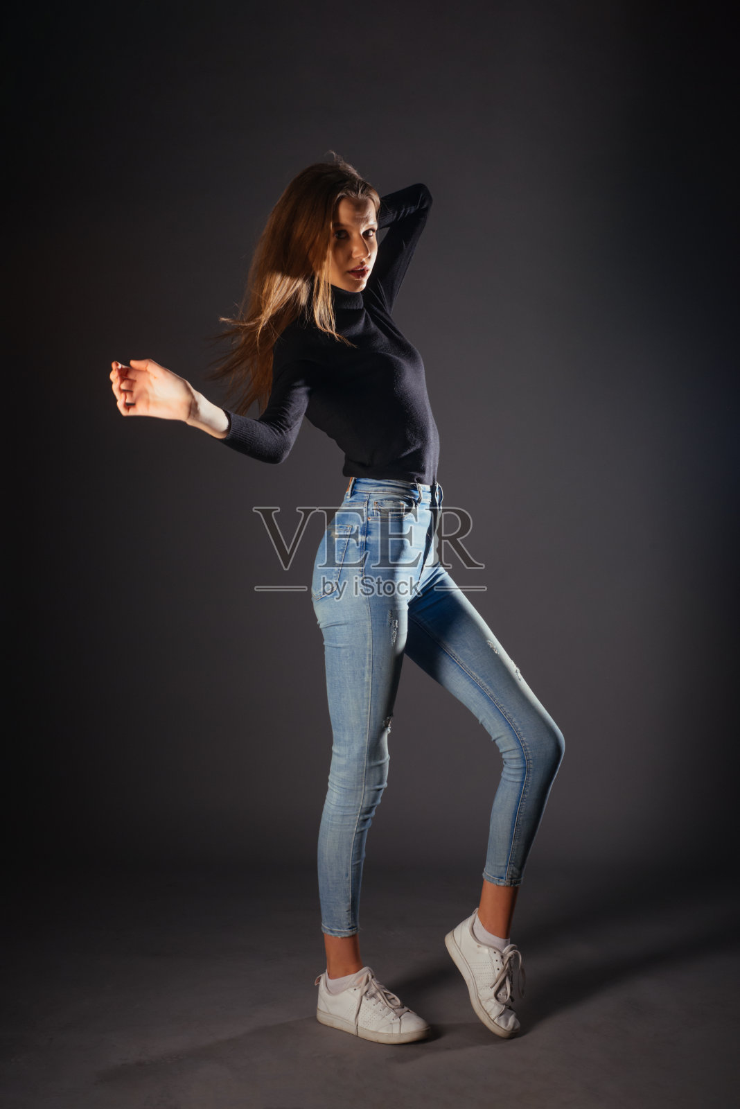 一个美丽的女模特穿着黑色的衬衫，牛仔裤和白色的运动鞋在工作室的黑色背景摆姿势的全长照片照片摄影图片