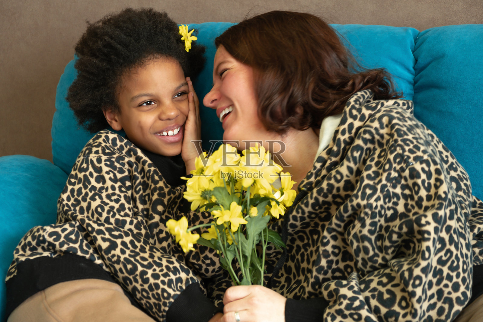 一位白人母亲和一位非裔美国人的女儿一起坐在家里的沙发上，拥抱着，笑着。女孩给了她妈妈黄花。生日快乐，母亲节祝福。照片摄影图片