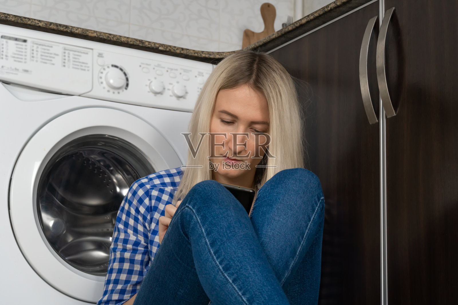 近距离观察年轻的金发女人，严肃地看着她的手机，坐在厨房洗衣机旁边照片摄影图片