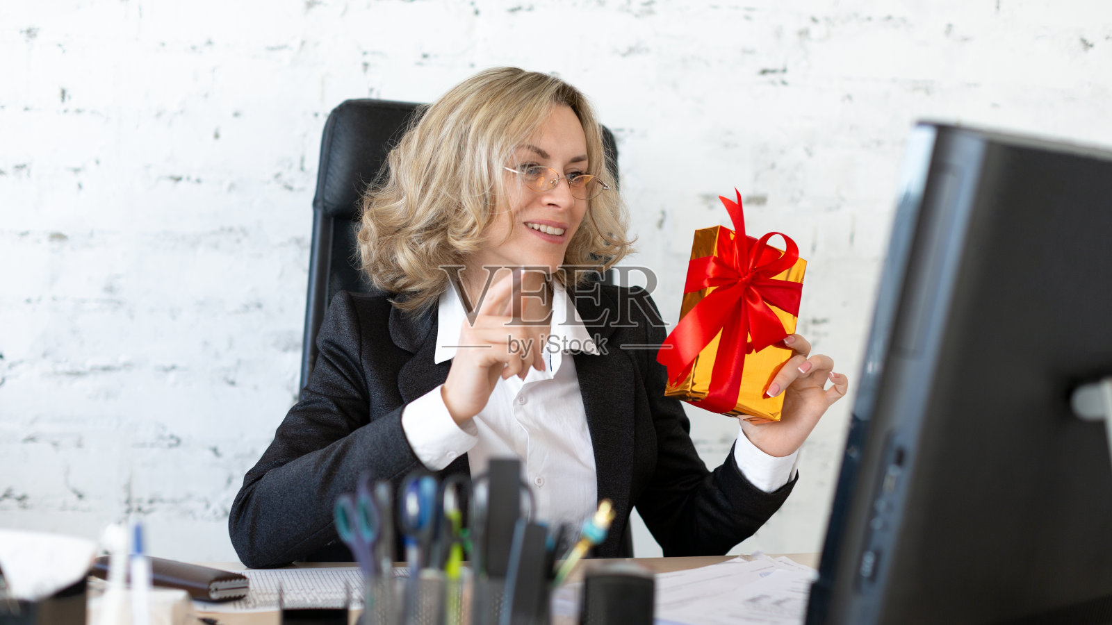 在工作场所给自己一个惊喜。快乐的成年女子穿着正式的衣服，手里拿着一个红色的礼品盒放在办公室的办公桌上，对着电脑屏幕，选择性的专注。照片摄影图片