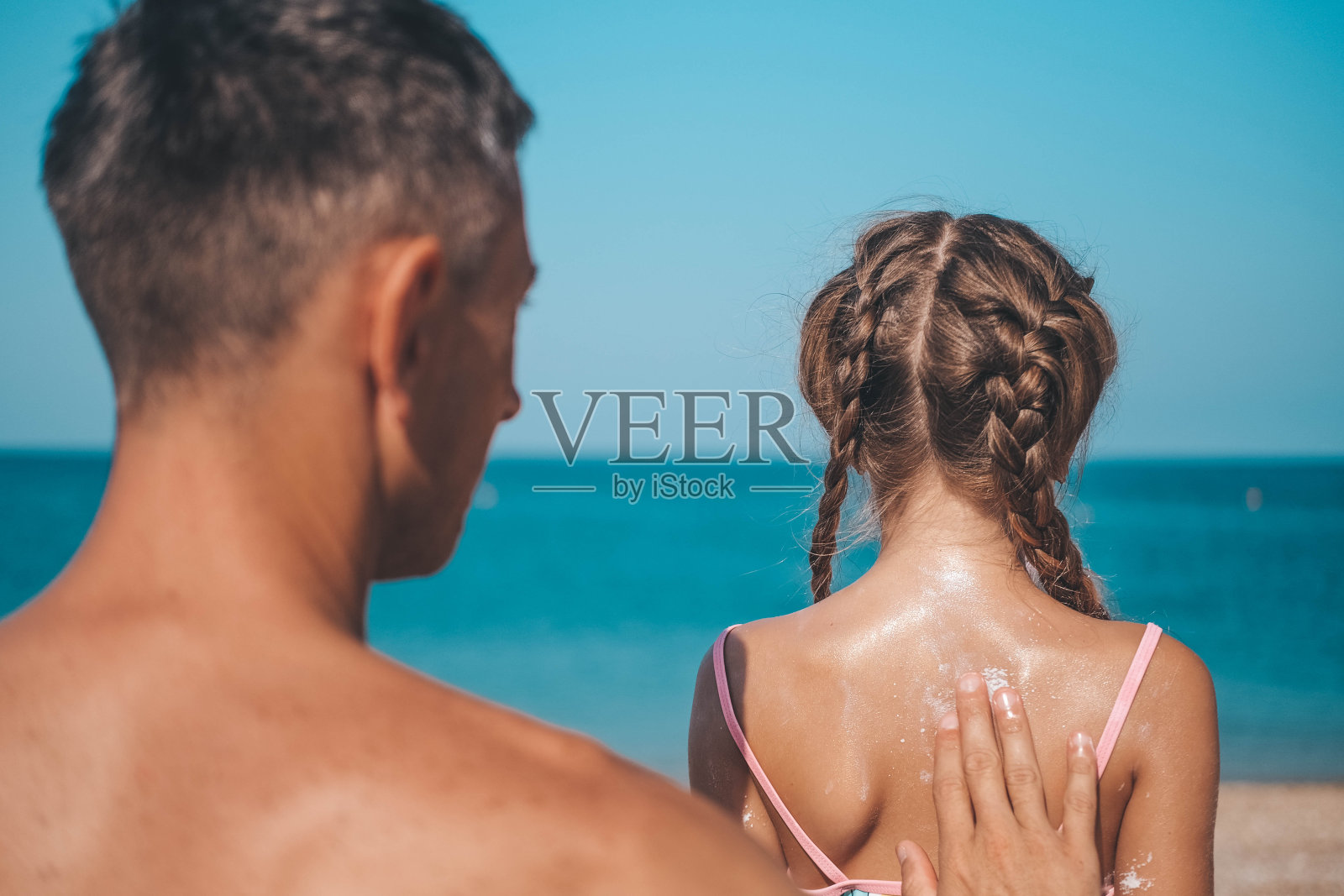一位父亲在沙滩上给女儿的背部涂护肤霜。一个男人把防晒霜涂抹在一个孩子的身上。海边涂着防晒霜的可爱小女孩。副本的空间。照片摄影图片