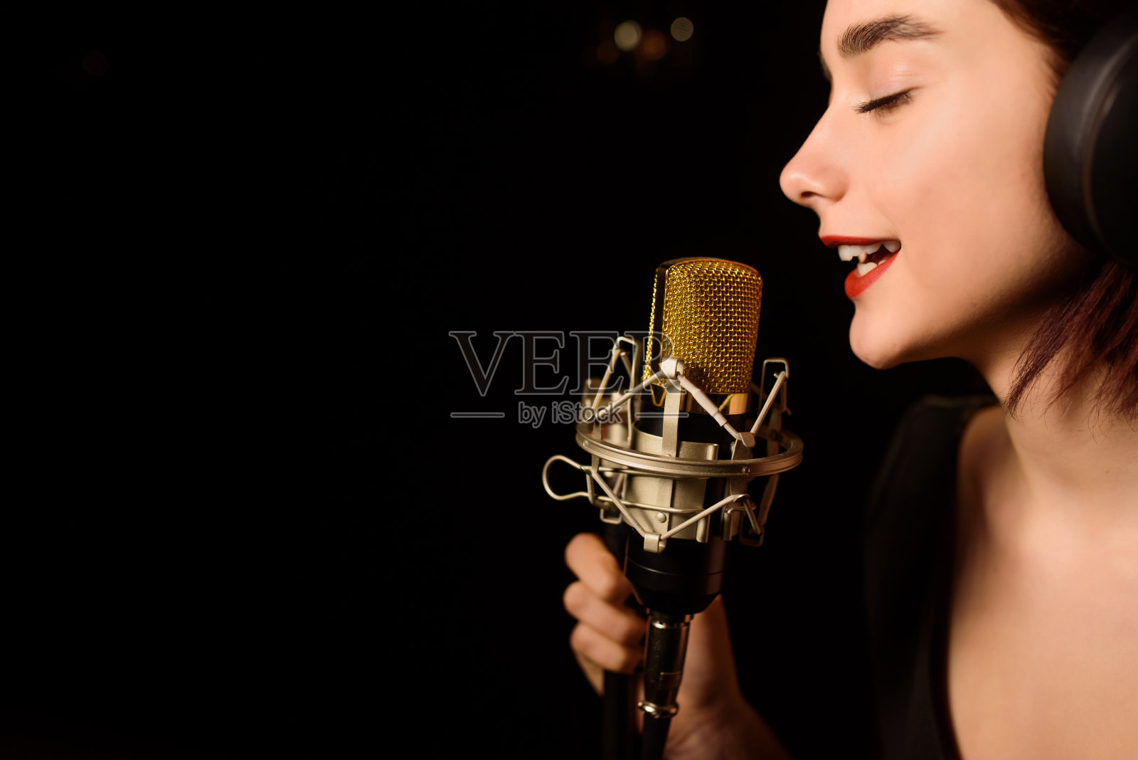 一名戴着耳机的年轻女子正在录音室里用黑色背景录制歌曲。照片摄影图片