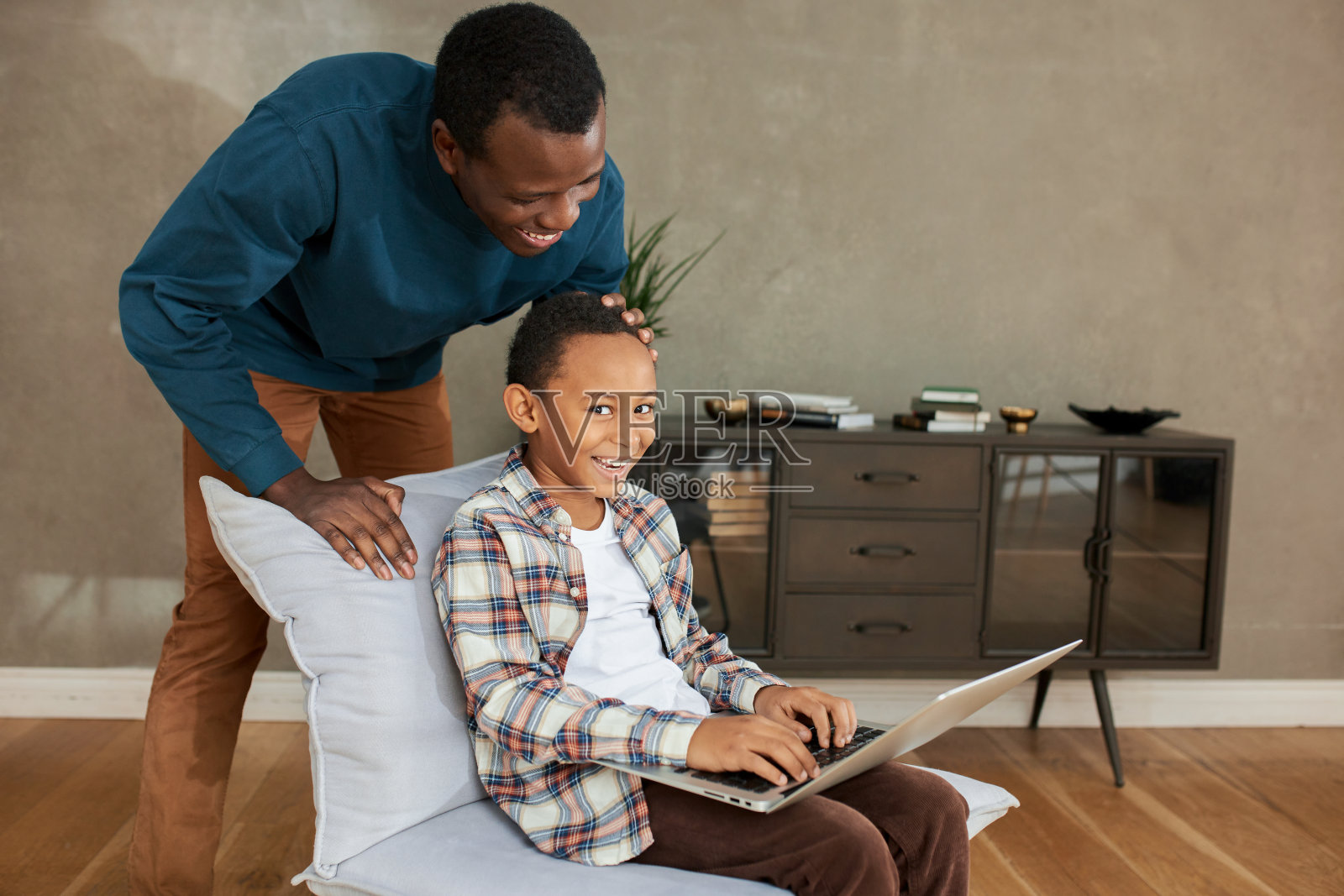这是一张有趣的照片，一个深色皮肤的卷发小男孩笑着把笔记本电脑放在膝盖上，坐在舒适的椅子上，被他英俊的父亲惊喜地拍着他的头，微笑着照片摄影图片