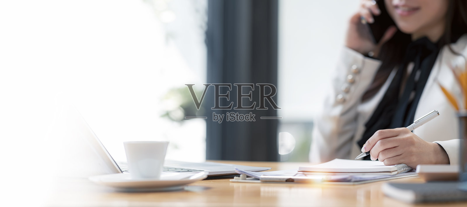 在现代的办公室里，一名穿着西装的年轻女子坐在办公桌前，用手机、笔记本电脑思考和撰写商业计划。房间里,复制空间。照片摄影图片