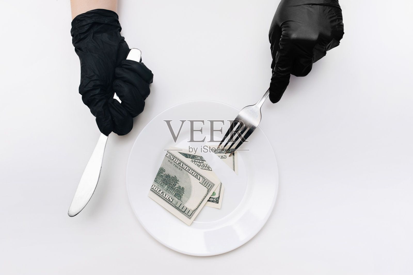 商务午餐的概念。戴着黑色手套的女性手拿着刀叉，从一张百元大钞上切下一块放在一个白色盘子上。设想可视化税收，贬值和利息。照片摄影图片