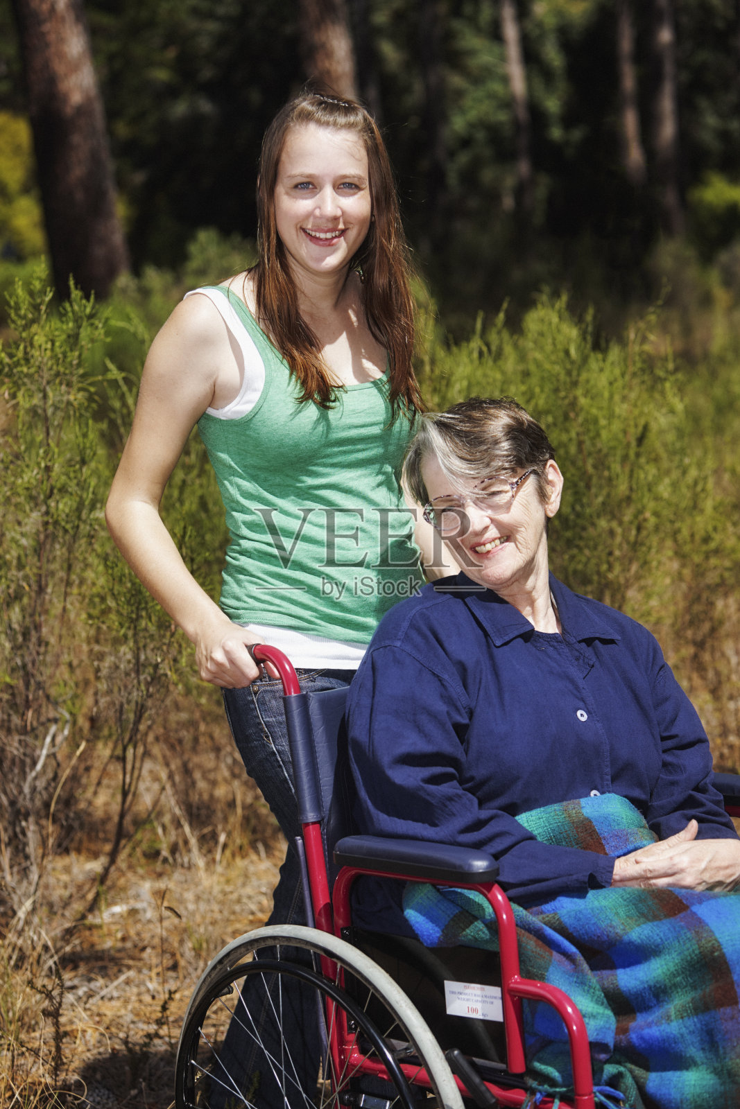 坐在轮椅上的老年妇女和年轻的亲戚或照顾者，在阳光明媚的树林里照片摄影图片