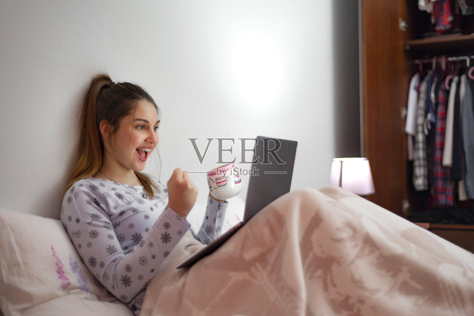 漂亮的年轻女孩躺在床上看着她的笔记本电脑看到她赢了，表达了她的兴奋。照片摄影图片