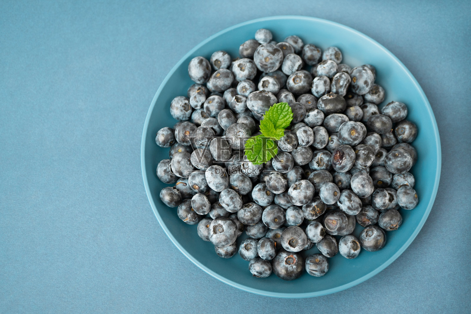 蓝色的背景上放着一个装满成熟蓝莓的蓝色碗。碗上装饰着新鲜的薄荷叶。适当的营养,维生素。素食的概念。浆果。俯视图照片摄影图片