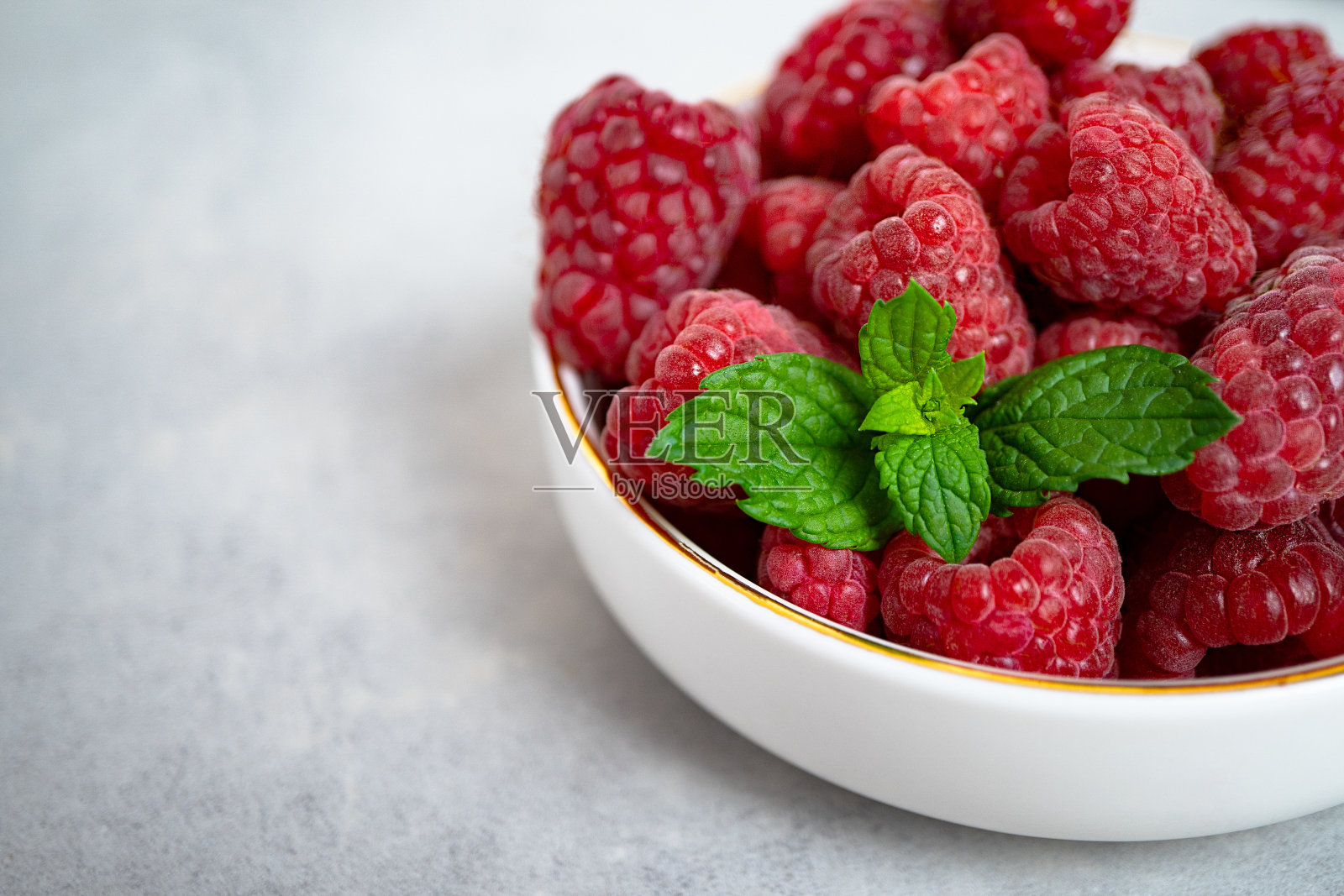 甜熟的覆盆子和薄荷叶放在一个白色的碗里，边缘是金色的，站在灰色的混凝土背景上。健康的甜点，适当的营养。季节性的新鲜浆果。特写镜头照片摄影图片