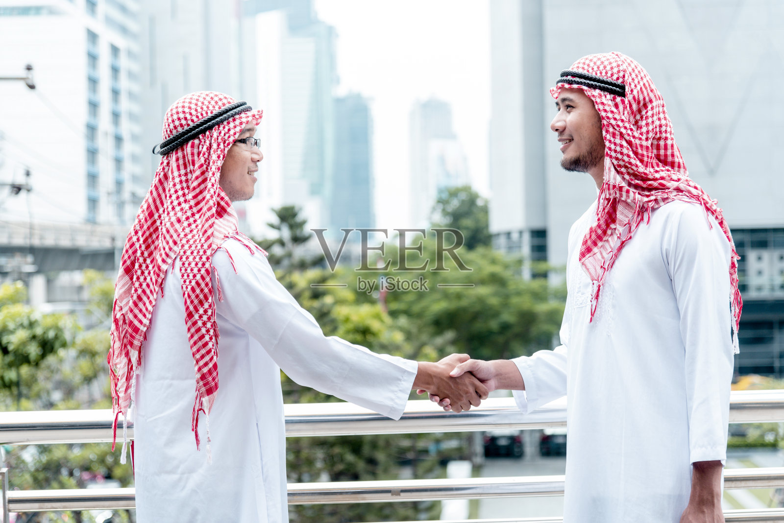 穿着穆斯林服装的阿拉伯商人，在阿联酋现代城市背景下握手。穆斯林团队合作的商业伙伴通过合作握手，多元种族阿拉伯人信任承诺照片摄影图片