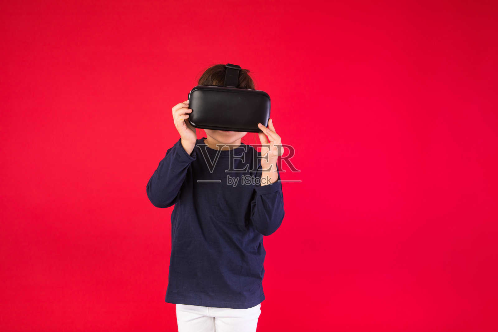 前面的小女孩戴着虚拟现实眼镜，在红色的背景上用她的手虚拟地触摸。虚拟、增强现实、技术、游戏和童年概念。照片摄影图片