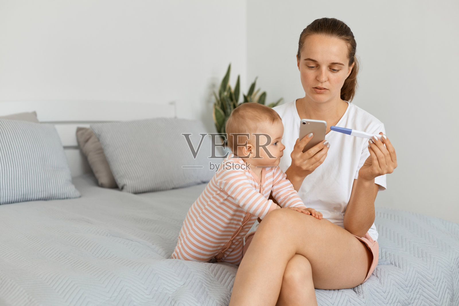 身穿白色t恤和短裤的黑头发的妇女抱着验孕棒，坐在床上，拨通了电话。照片摄影图片