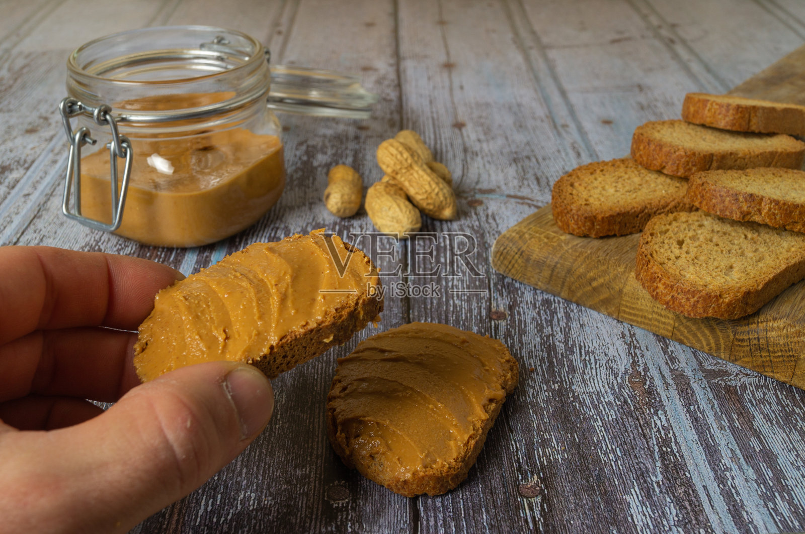 一个男人抓着涂满花生酱的吐司的手的特写镜头，装满花生酱的玻璃容器，烤面包的木板，自制的花生酱，自然的，在一个质朴的木制底座上。照片摄影图片