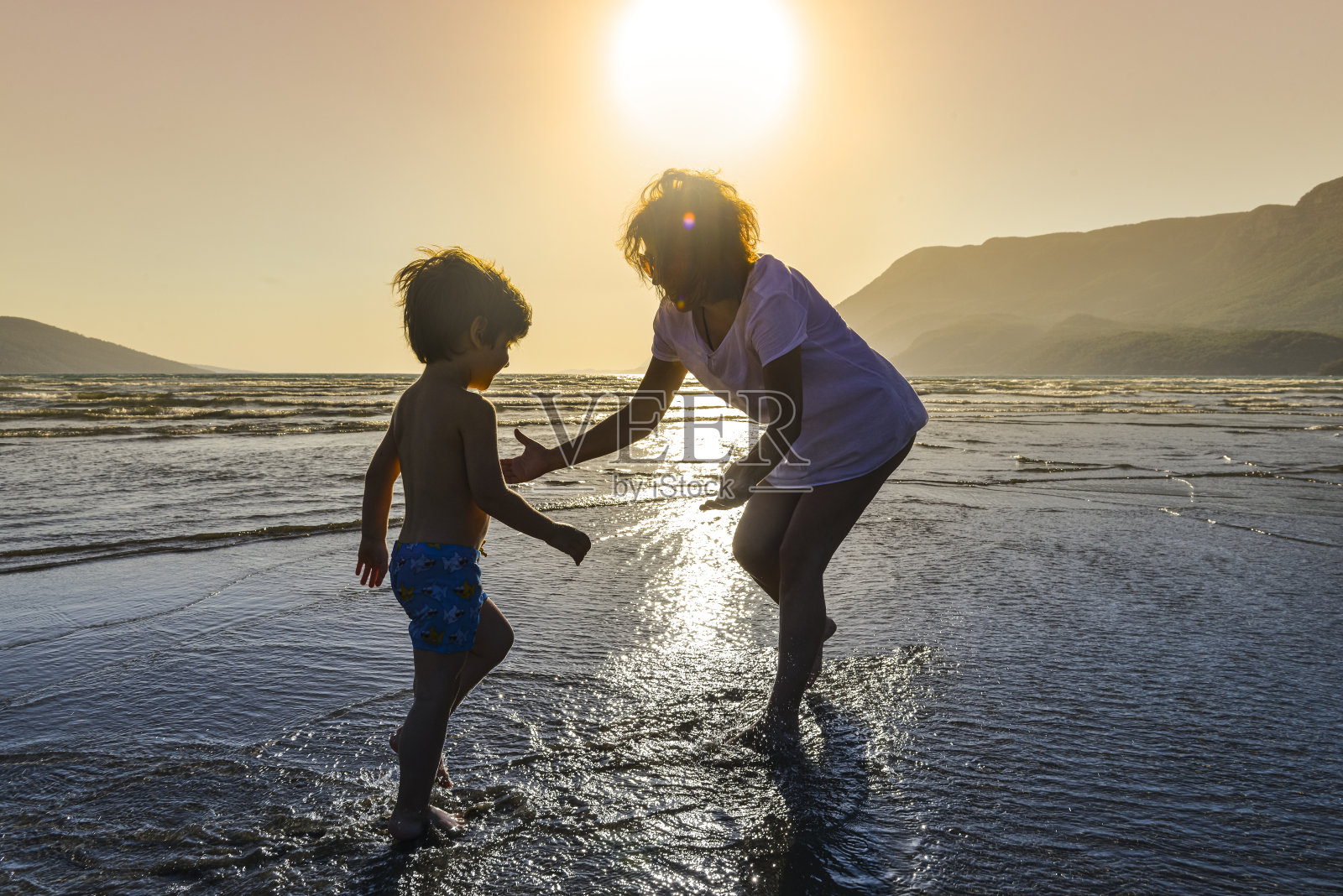 夕阳西下的沙滩上，小男孩奔向妈妈的膝上。照片摄影图片