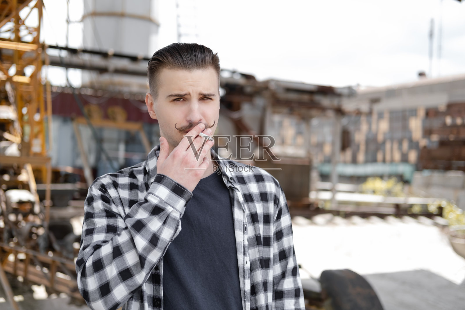 年轻英俊的男子，留着小胡子，穿着黑白格子衬衫，在外面抽烟。坏习惯照片摄影图片