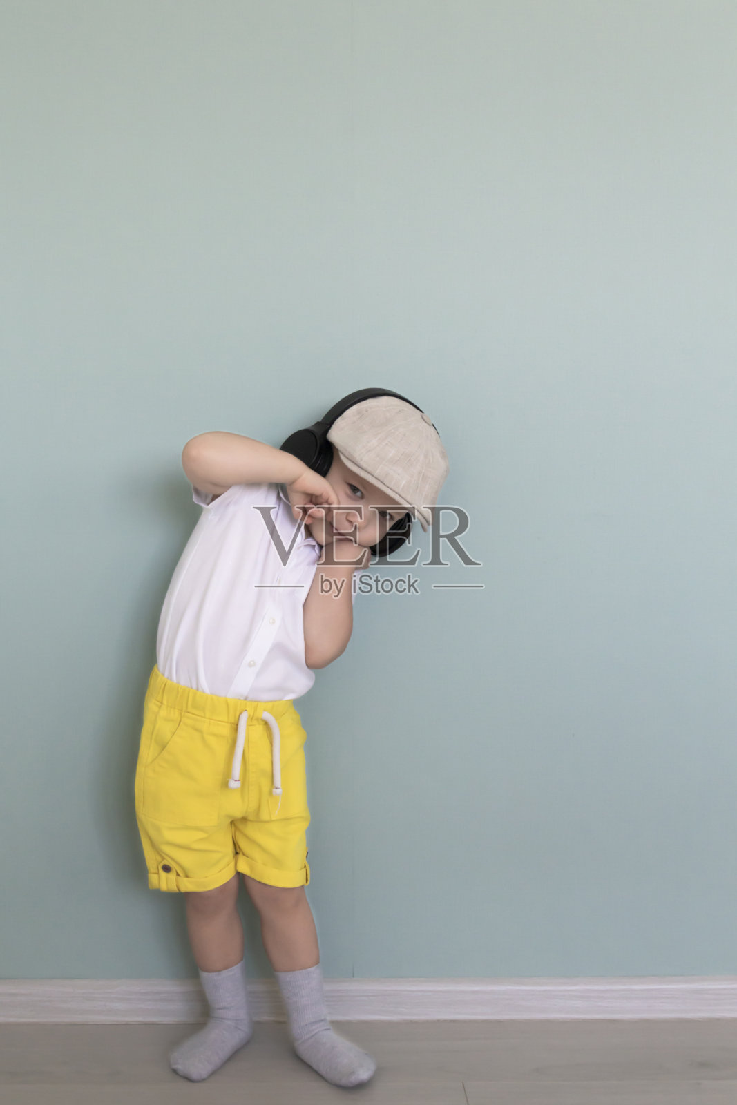 一个小男孩穿着白色衬衫，黄色短裤，戴着一顶复古的帽子，戴着大耳机听音乐。照片摄影图片