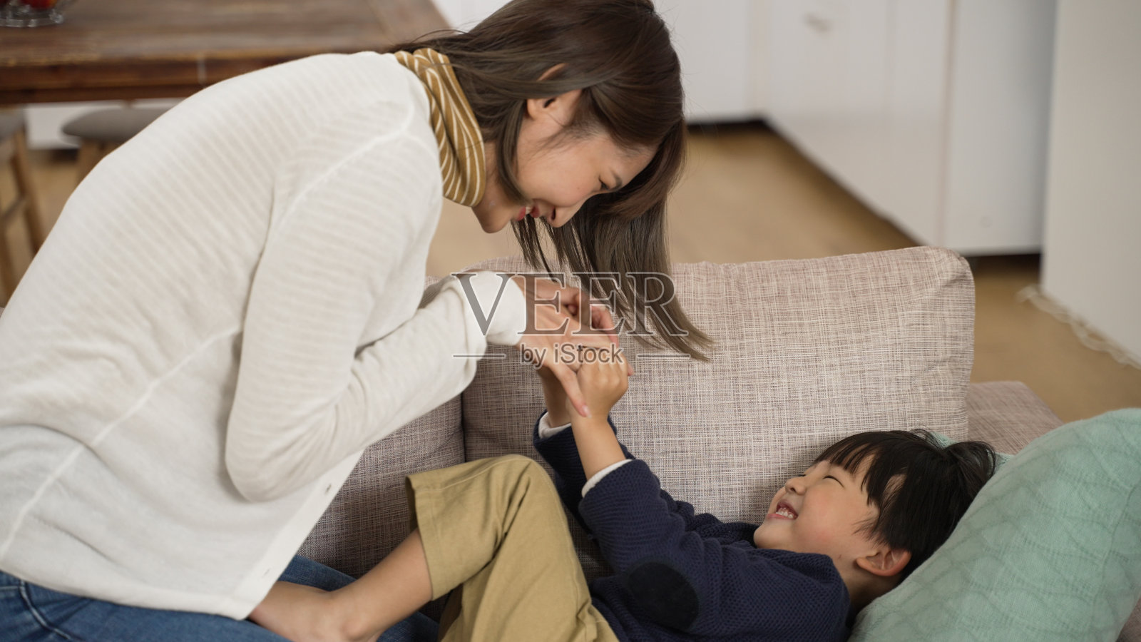 在家里客厅的沙发上，一个亚洲小男孩笑着试图把他妈妈推开，而他妈妈正在和他挠痒痒照片摄影图片