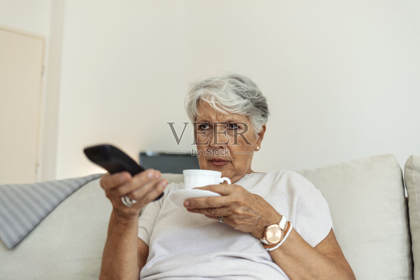 震惊的女人用电视遥控器坐在家里的沙发上。照片摄影图片