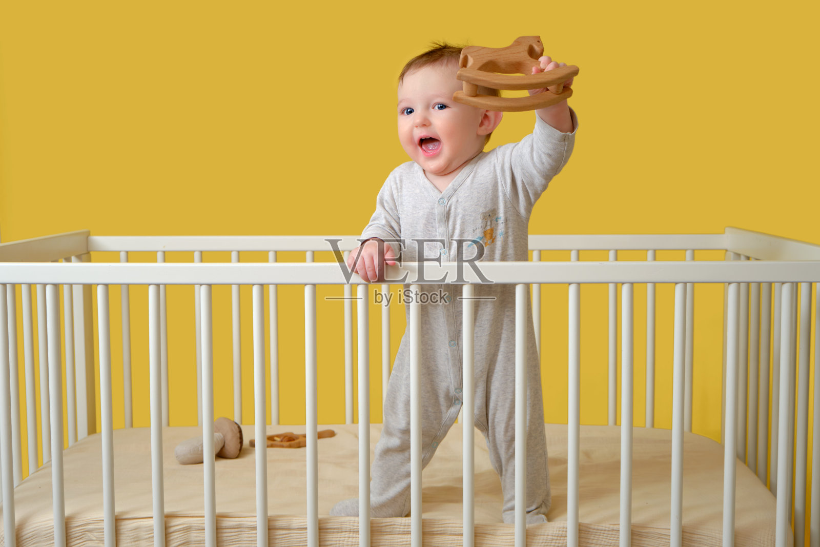 快乐的蹒跚学步的小男孩在婴儿床里，手里拿着一个木制玩具，黄色的画室背景。穿着睡衣微笑的孩子照片摄影图片