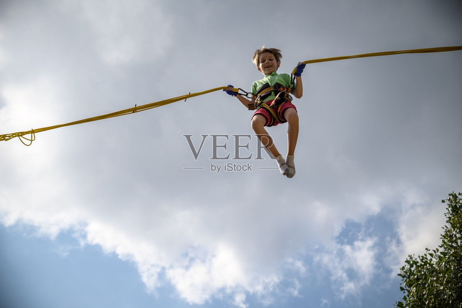 那个男孩正在蹦极蹦床上跳。一个带着保险和可伸缩橡皮筋的孩子悬挂在天空中。快乐童年的概念和游乐园的游戏照片摄影图片