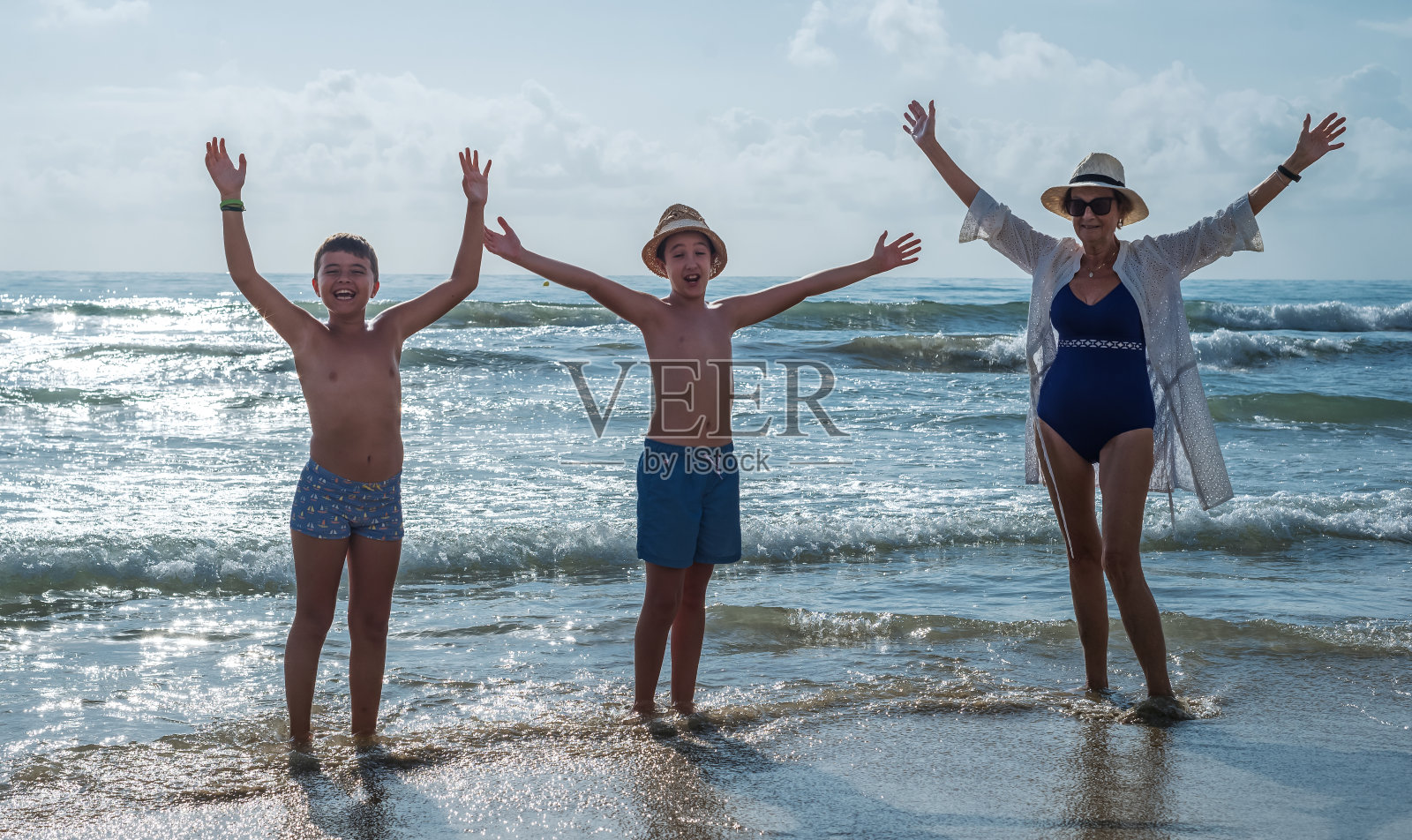 一位祖母和她的两个孙子在海滩上举起双臂。暑期家庭度假概念照片摄影图片