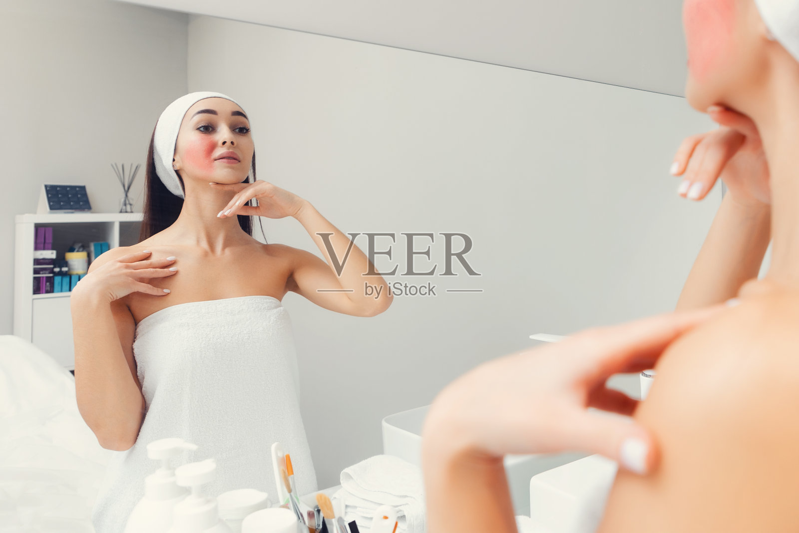 年轻美丽的女人与红血丝在她的脸颊上，站在浴室和看着镜子。酒渣鼻及护肤理念照片摄影图片