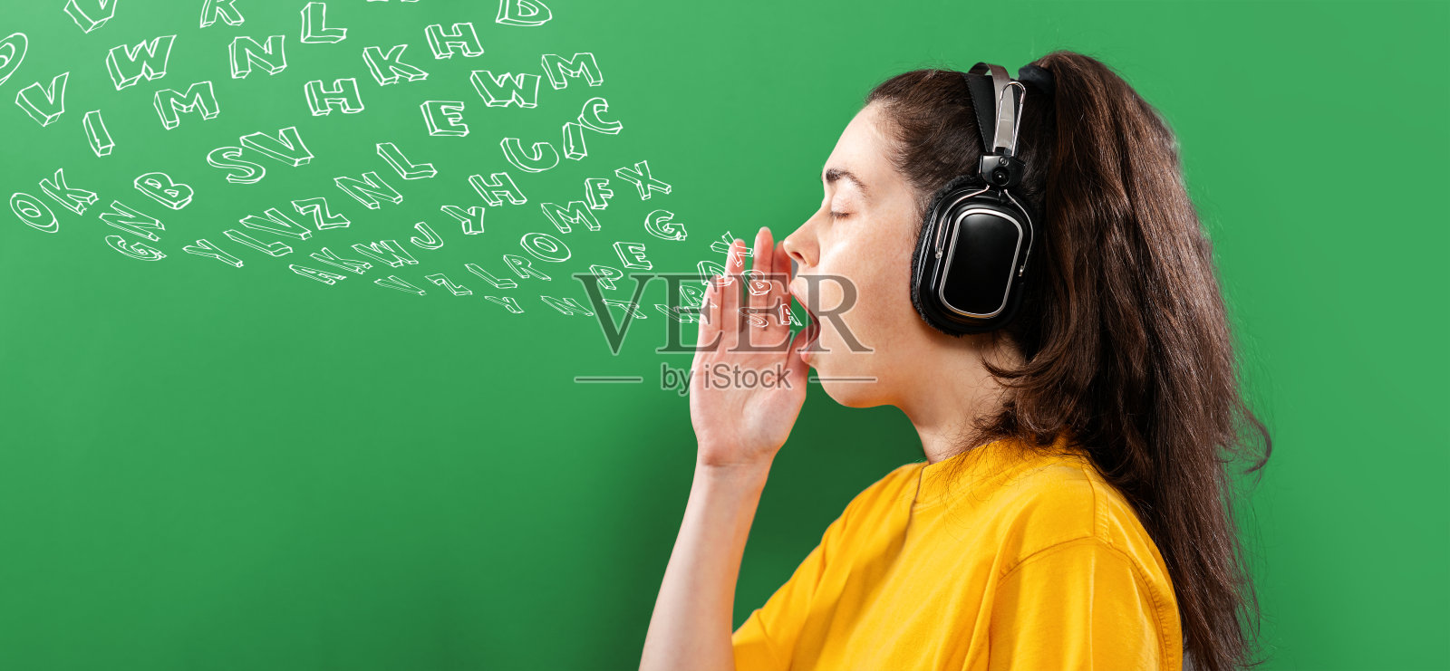 在线语言学习。一个戴着耳机的年轻女子的肖像，她的手捂着她张开的嘴。绿色背景与字母。侧视图。横幅。英语日的概念照片摄影图片