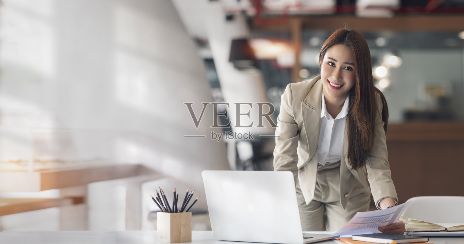 年轻美丽和迷人的亚洲女商人微笑着看着相机，而工作在笔记本电脑和文件在办公室。照片摄影图片