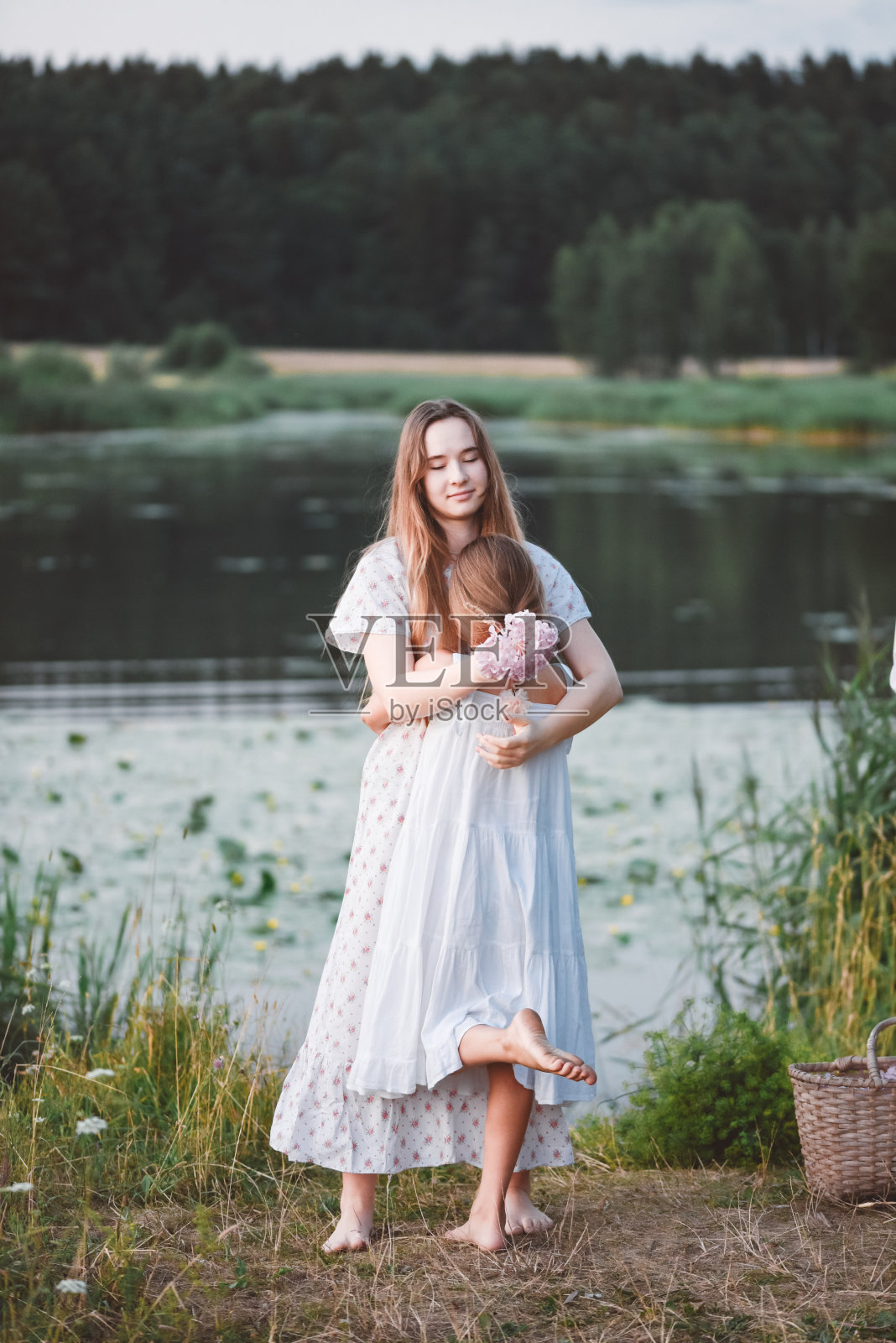 姐妹们拥抱着站在靠近森林的湖边。照片摄影图片