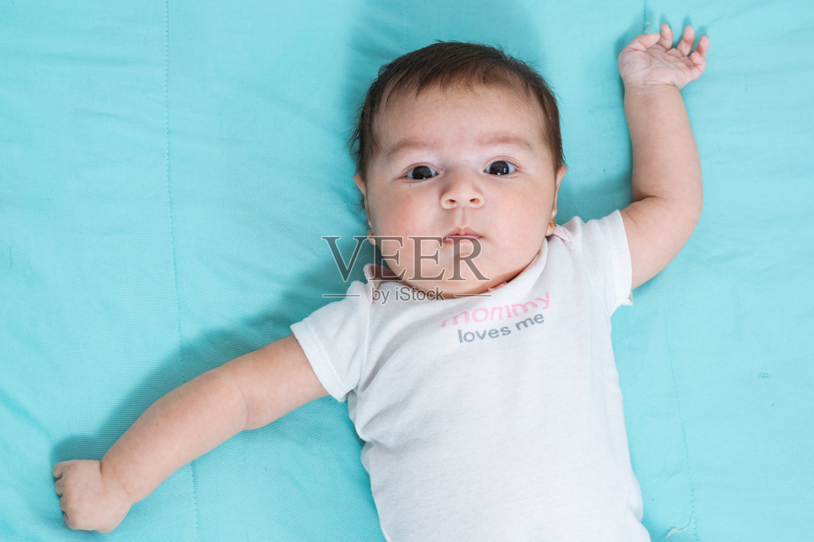 中拍美丽的两个月大的拉丁婴儿躺在蓝色的床单上，她的目光盯着相机，她的手在一个惊恐的姿势，黑色的眼睛，黑色的眼睛。照片摄影图片