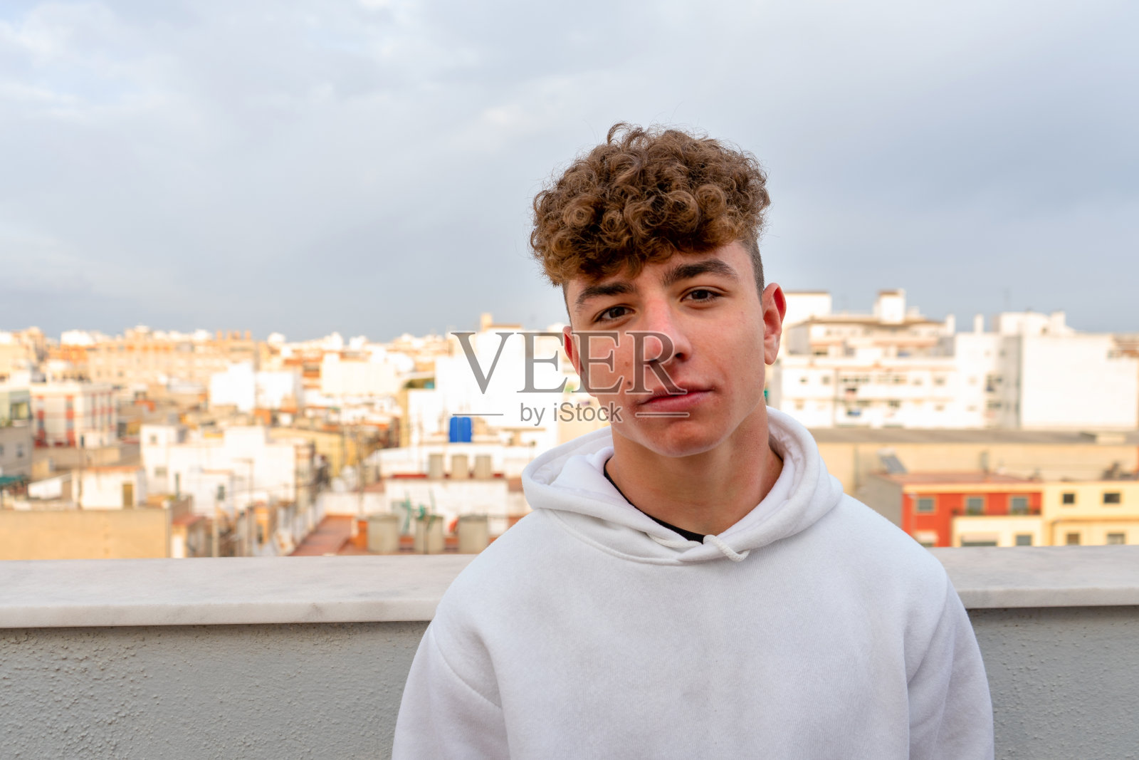 这是一幅严肃的年轻白人男子的肖像，头发卷曲，穿着白色运动衫，看着镜头，背景是城市的天际线。照片摄影图片