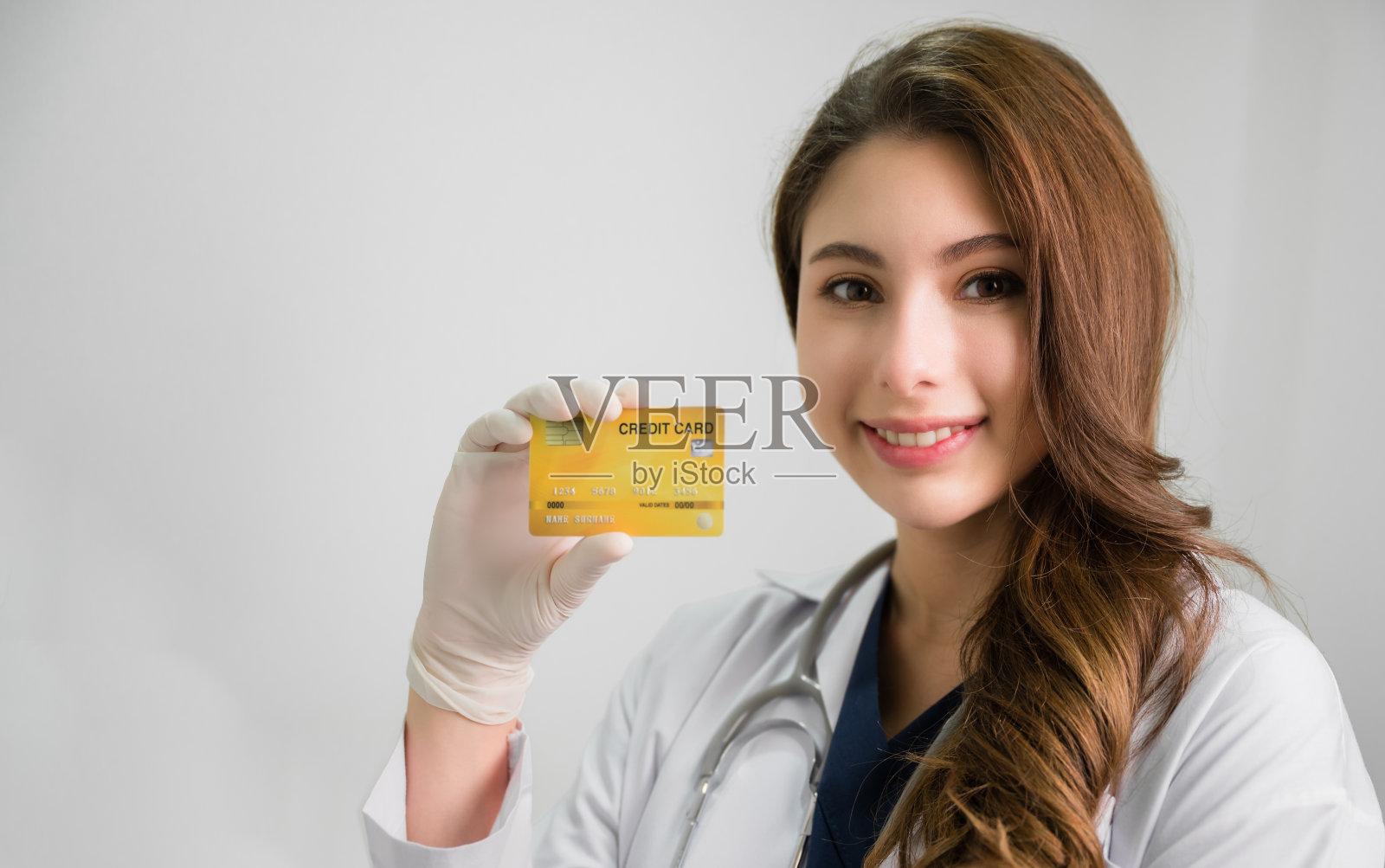 快乐美丽的亚洲女医生的肖像，穿着白大褂，站着听诊器，在医院的办公室里拿着信用卡。照片摄影图片