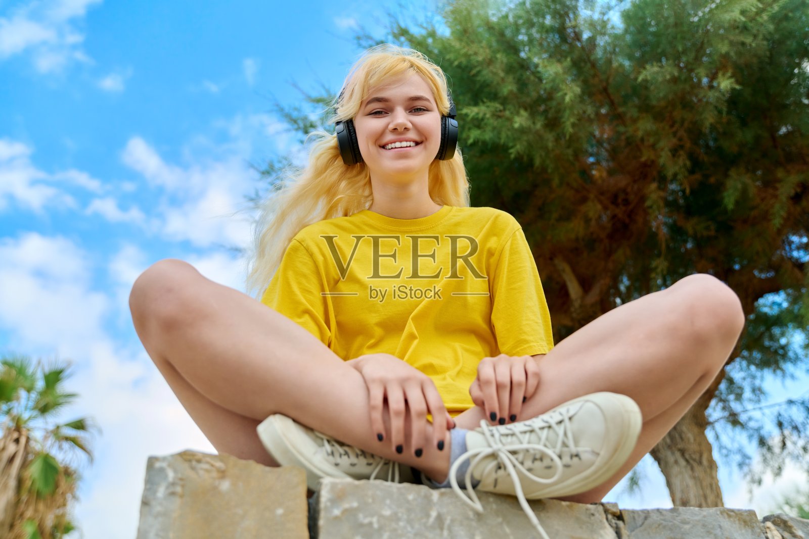 十几岁的女孩戴着耳机坐在公园的户外，蓝天白云照片摄影图片