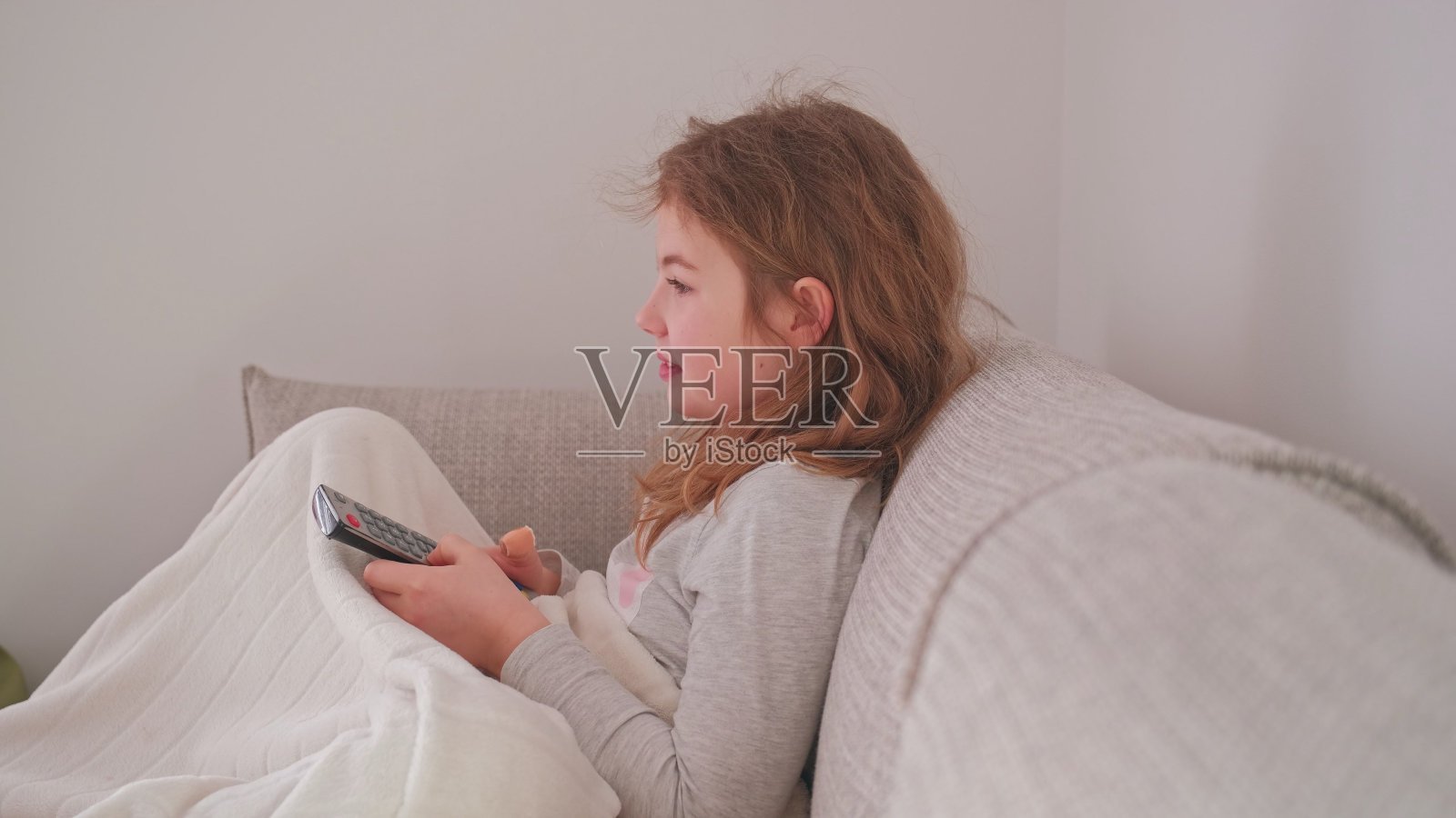 美丽的年轻微笑的白人女孩在睡衣坐在舒适的沙发在客厅看电视和按下按钮的遥控器在周末早上照片摄影图片