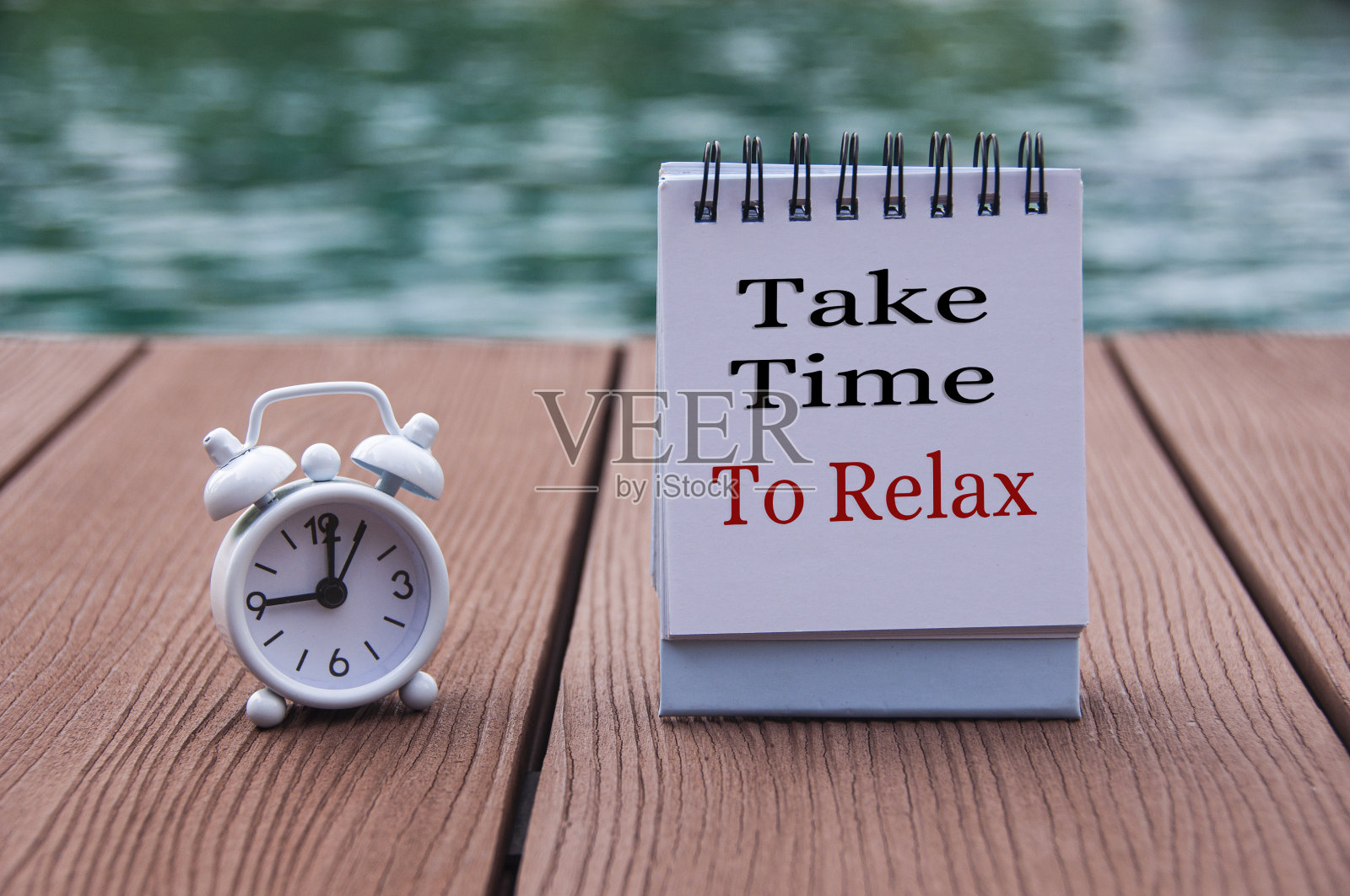 木地板上的闹钟和记事本上的文字——花点时间放松一下。与模糊的游泳池背景。钟定在九点钟。照片摄影图片