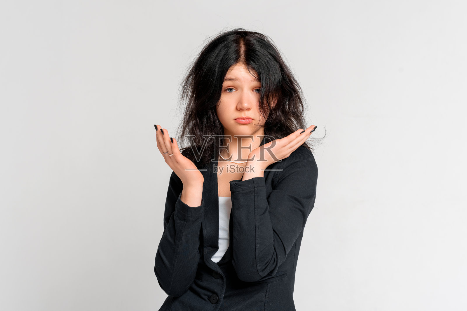 一个困惑的十几岁女孩的肖像，凌乱的头部，在黑色的运动夹克，摊开双手，看着相机与困惑的表情，站在灰色的背景照片摄影图片