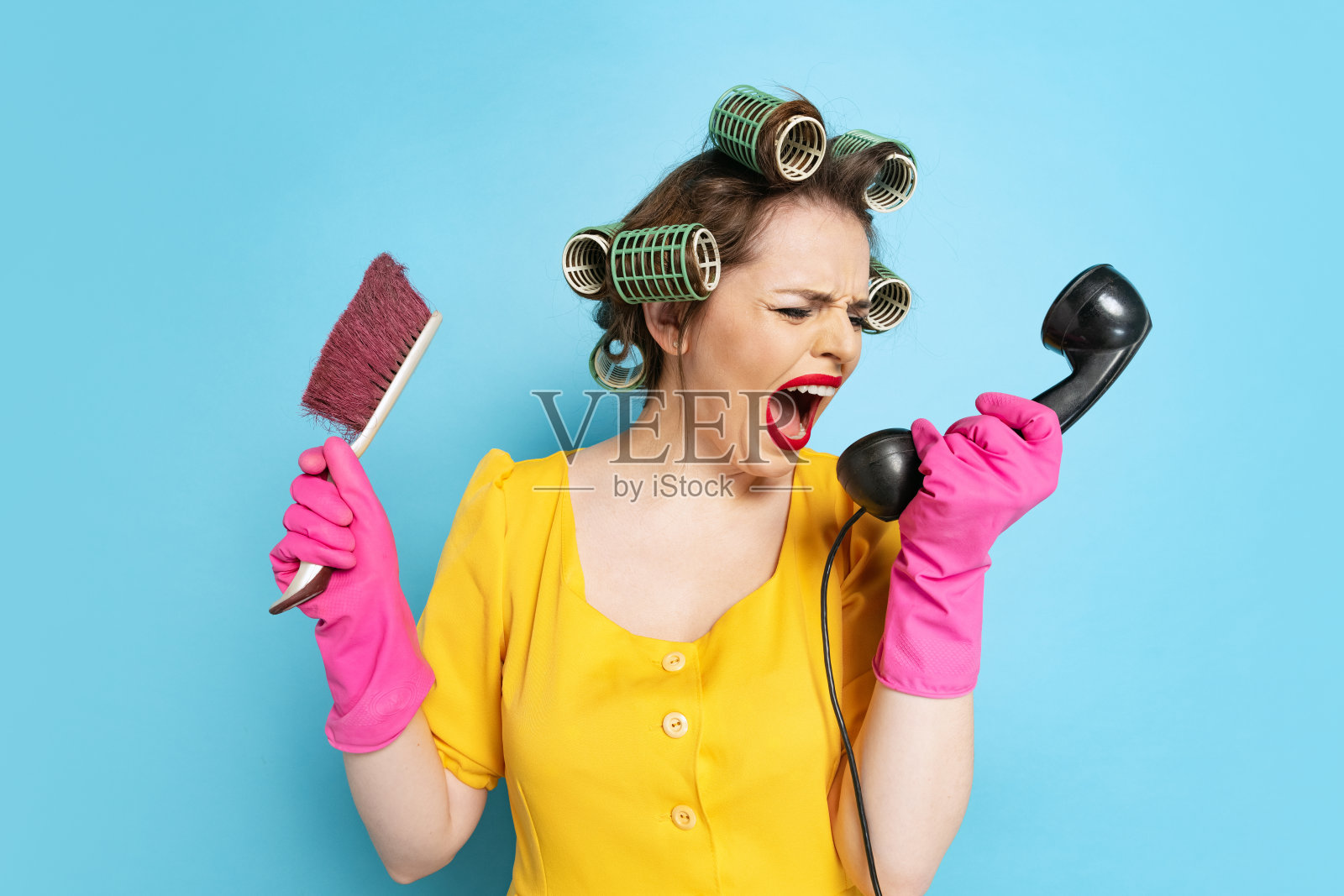 大吼大叫。年轻情绪化的女孩，家庭主妇，头上戴着卷发器，在蓝色工作室背景下用复古手机通话。情感，女性权利，美丽，家庭照片摄影图片