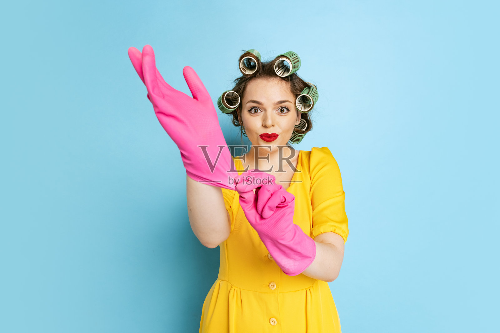 震惊的年轻漂亮的女孩，家庭主妇与卷发器在她的头孤立在蓝色工作室背景。情感，女性权利，美丽，家庭照片摄影图片