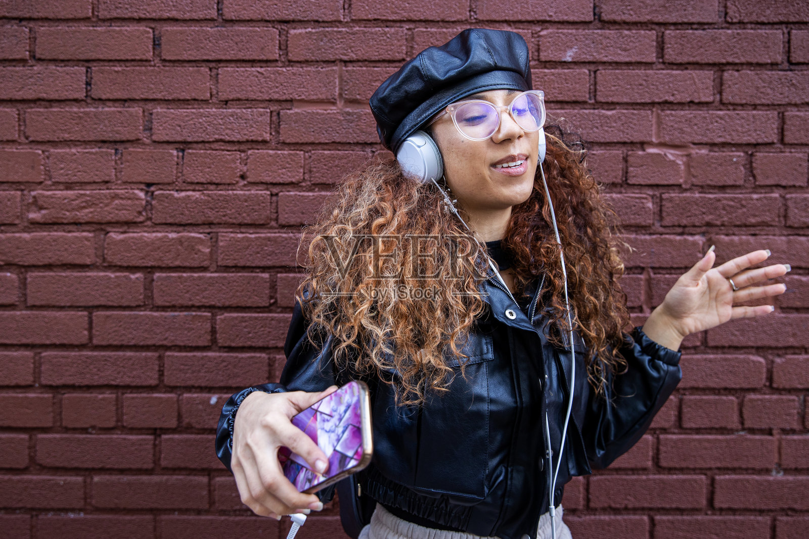 年轻女子穿着黑色皮夹克，戴着贝雷帽，边听音乐边在砖墙边跳舞照片摄影图片
