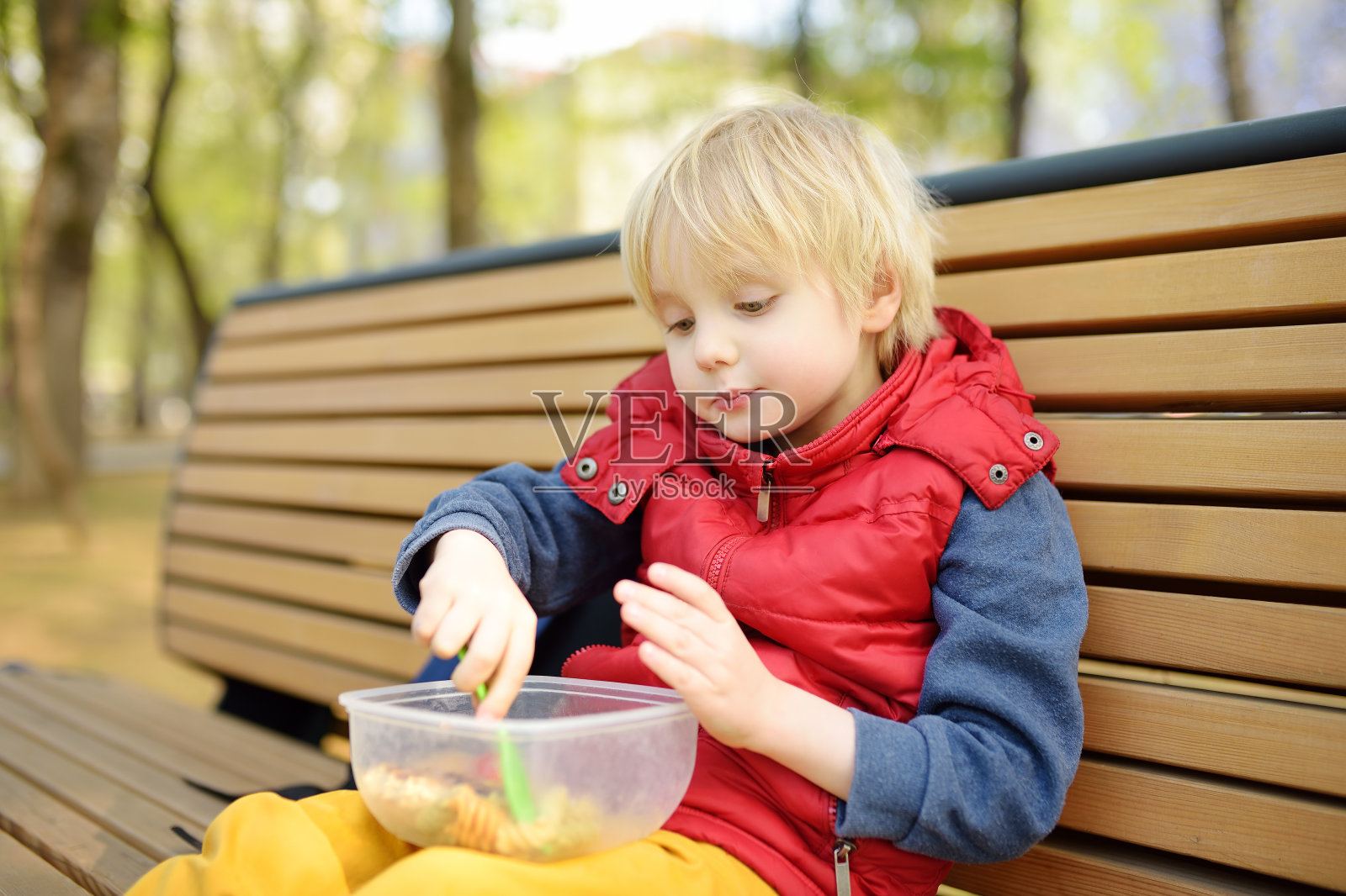 幼儿园或学校放学后，一个小男孩坐在公园长凳上的塑料容器里吃午饭。在街上为孩子们带走食物。给孩子们的健康食品。照片摄影图片