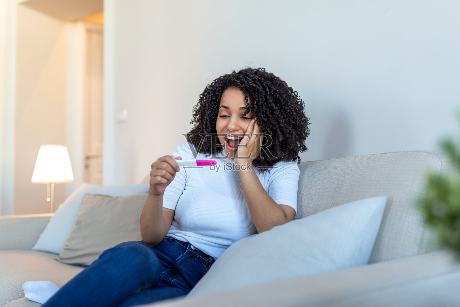 一个微笑的女人坐在沙发上，拿着阳性的怀孕检测结果，用她的智能手机和父亲聊天，告诉他一个好消息。微笑的怀孕女孩在手机上告诉好消息。照片摄影图片