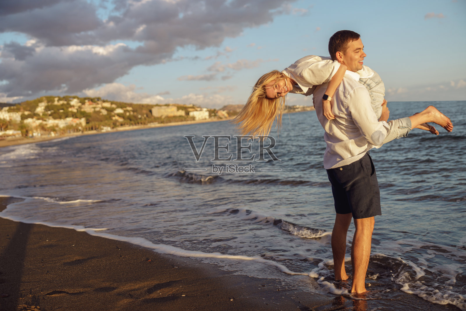 强壮的男人抱着他的女朋友的肩膀，站在沙滩上接近到来的海浪照片摄影图片