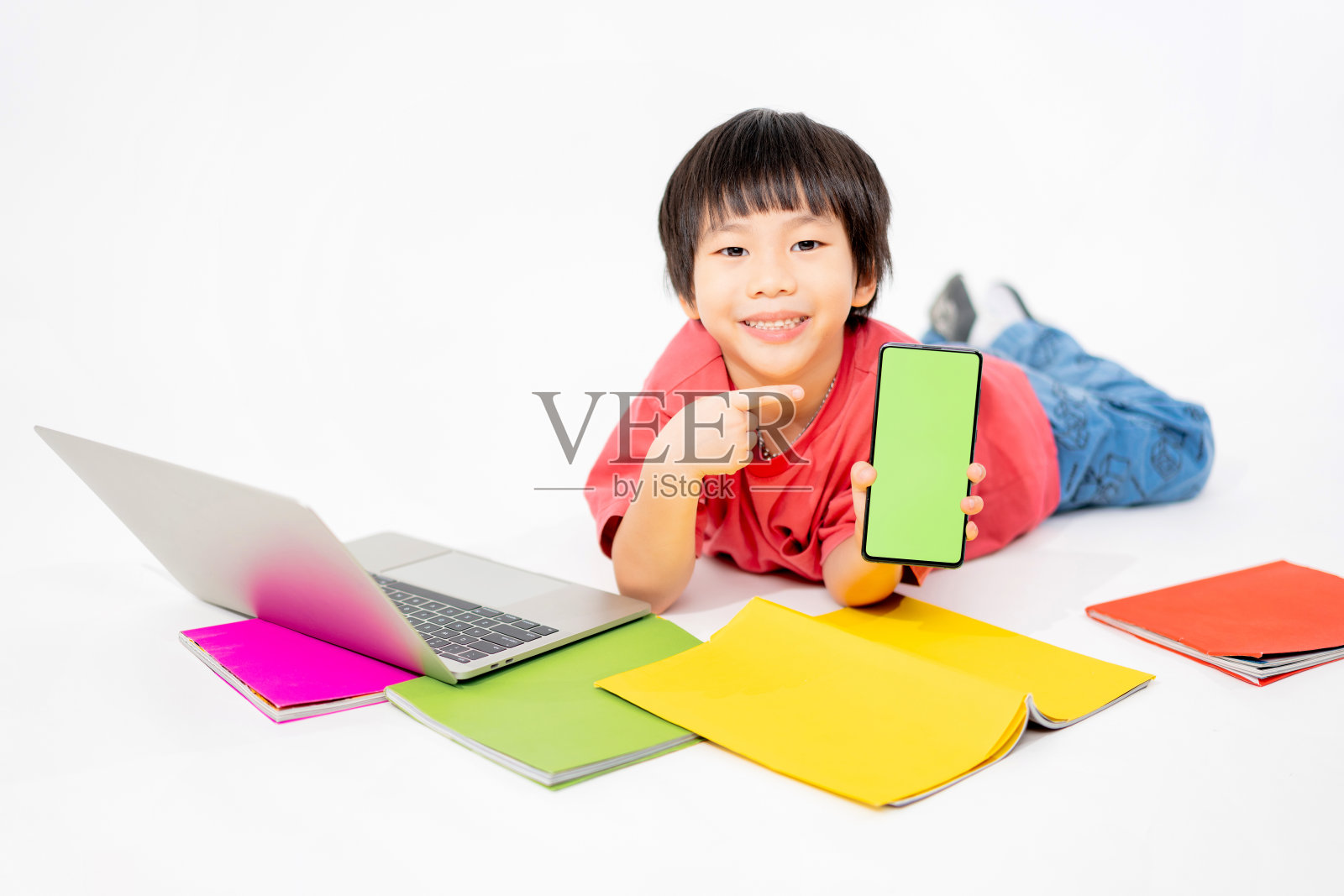 快乐亚洲男孩的肖像握着手机在他的手与地板上的绿色屏幕与笔记本电脑和书籍隔离在白色背景与复制空间，教育和学习与技术概念照片摄影图片