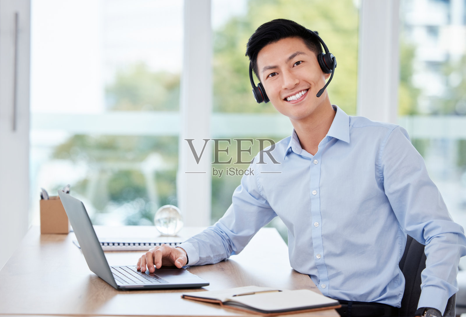 这张照片拍摄的是一名年轻的男性呼叫中心代理在工作时使用笔记本电脑并在笔记本上写字照片摄影图片