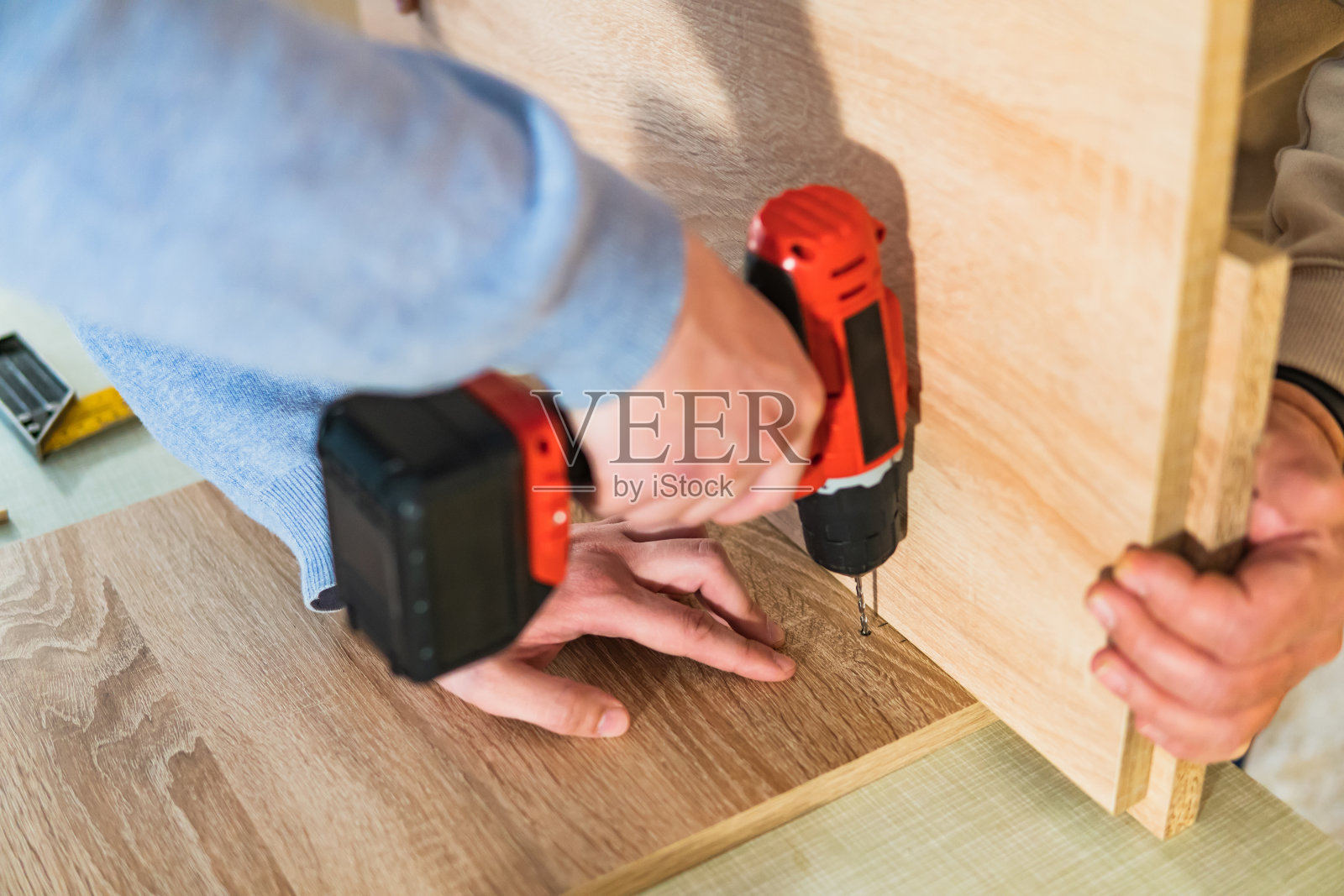 一个年轻人的手的特写，拿着一个钻头和钻一个木板(胶合板)，而另一个人拿着另一块木板。在家DIY项目。照片摄影图片