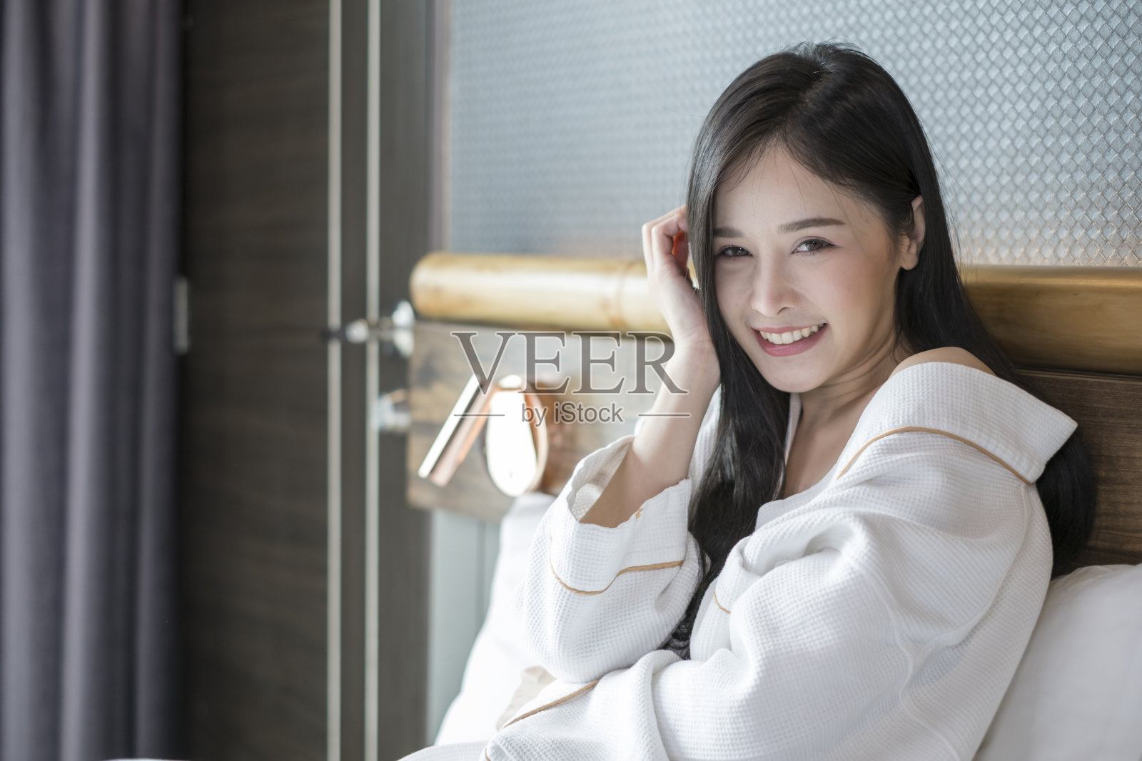 快乐美丽的年轻亚洲女人放松在床上早上。年轻漂亮的女人穿着白色浴衣在舒适的卧室。照片摄影图片