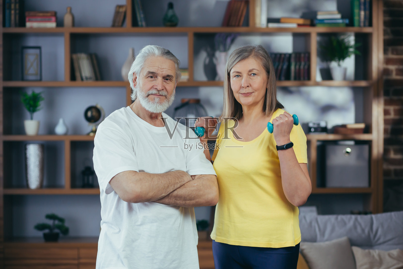 退休老人夫妇拿着哑铃站在客厅里，微笑着看着镜头，正在健身照片摄影图片